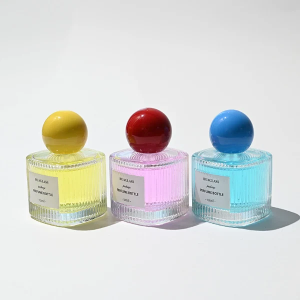 HUA0203 perfume bottle, cylindrical ribbed transparent glass body with thick base, 50 ml or 100 ml liquid, spherical glossy plastic cap in red or yellow, brand label applied on the surface, modern luxurious design, ideal for gifting and daily use.