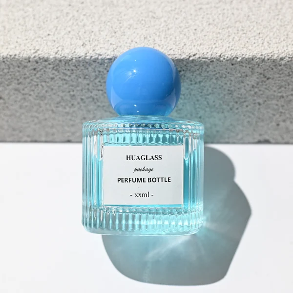 HUA0203 perfume bottle, cylindrical ribbed transparent glass body with thick base, 50 ml or 100 ml liquid, spherical glossy plastic cap in red or yellow, brand label applied on the surface, modern luxurious design, ideal for gifting and daily use.