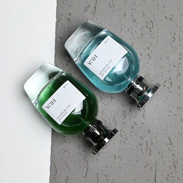 HUA0204 perfume bottle, flattened rounded streamlined transparent glass body with thick base and high-gloss finish, 100 ml liquid, flat cylindrical Zamac cap with slightly enlarged flat top fitting snugly on the neck, printed logo label, modern luxurious design, ideal for gifting and daily use.