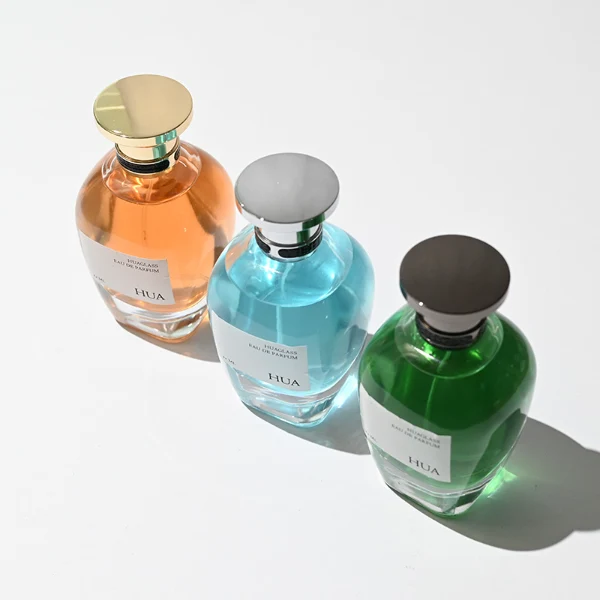 HUA0204 perfume bottle, flattened rounded streamlined transparent glass body with thick base and high-gloss finish, 100 ml liquid, flat cylindrical Zamac cap with slightly enlarged flat top fitting snugly on the neck, printed logo label, modern luxurious design, ideal for gifting and daily use.