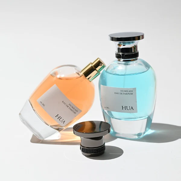 HUA0204 perfume bottle, flattened rounded streamlined transparent glass body with thick base and high-gloss finish, 100 ml liquid, flat cylindrical Zamac cap with slightly enlarged flat top fitting snugly on the neck, printed logo label, modern luxurious design, ideal for gifting and daily use.