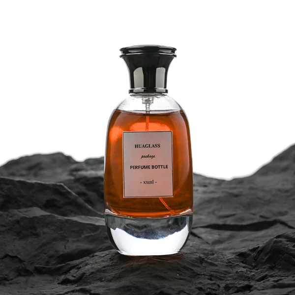 HUA0205 perfume bottle, flattened rounded streamlined transparent glass body with thick base and high-gloss finish, 100 ml liquid, flat cylindrical Zamac cap in black, gold, or silver with slightly enlarged flat top fitting snugly on the neck, brand label applied on the surface, modern luxurious design, ideal for gifting and daily use.
