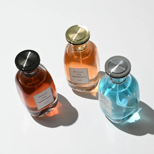 HUA0205 perfume bottle, flattened rounded streamlined transparent glass body with thick base and high-gloss finish, 100 ml liquid, flat cylindrical Zamac cap in black, gold, or silver with slightly enlarged flat top fitting snugly on the neck, brand label applied on the surface, modern luxurious design, ideal for gifting and daily use.