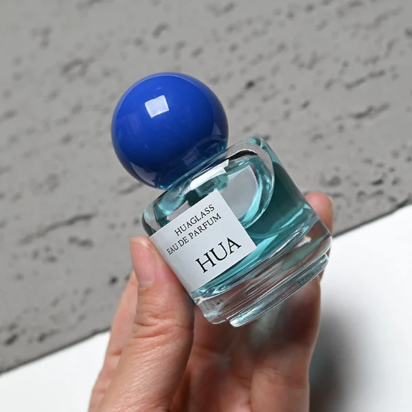 HUA0208 perfume bottle, cylindrical transparent glass body with thick base, 30 ml liquid, spherical plastic cap in blue, red, or yellow, brand label applied on the surface, modern playful luxury design, ideal for gifting and daily use.