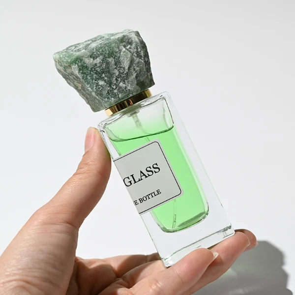 HUA0213 perfume bottle, rectangular transparent glass body with thick base, 30 ml, 50 ml, or 100 ml liquid, rough natural crystal stone cap with gold neck ring, brand label applied on the bottle surface, modern artistic luxury design, ideal for gifting and daily use.