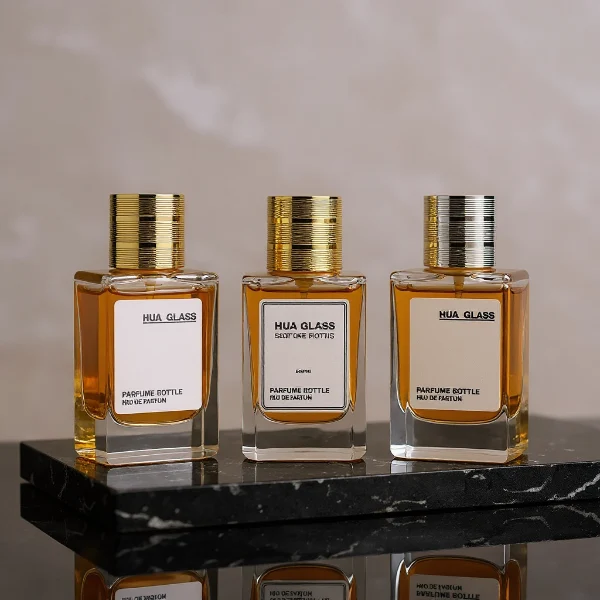HUA0235 perfume bottle, square transparent glass body with thick base, 30 ml, 50 ml, or 100 ml liquid, cylindrical plastic cap with side threading and metallic finish in gold, black, or silver, brand label applied on bottle, modern luxurious design, ideal for gifting and daily use.
