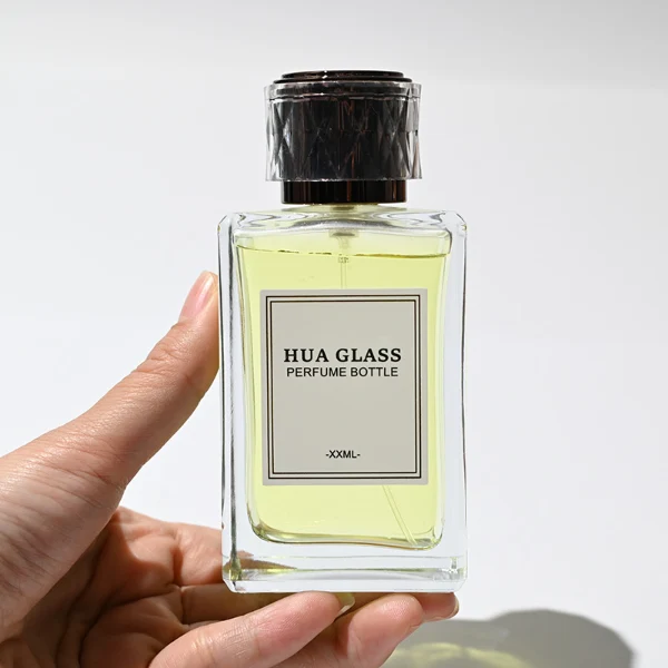 HUA0250 perfume bottle, rectangular transparent glass body with thick base, 100 ml liquid, black cylindrical plastic inner cap with transparent outer plastic, brand label applied on bottle, modern luxurious design, ideal for gifting and daily use.