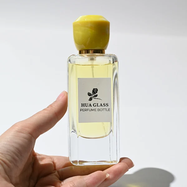 HUA0252 perfume bottle, rectangular transparent glass body with rounded edges and thick base, 50 ml or 80 ml liquid, yellow or red faceted geometric resin cap with smooth glossy finish, gold-colored bottle neck, brand label applied on bottle, modern artistic luxury design, ideal for gifting and daily use.