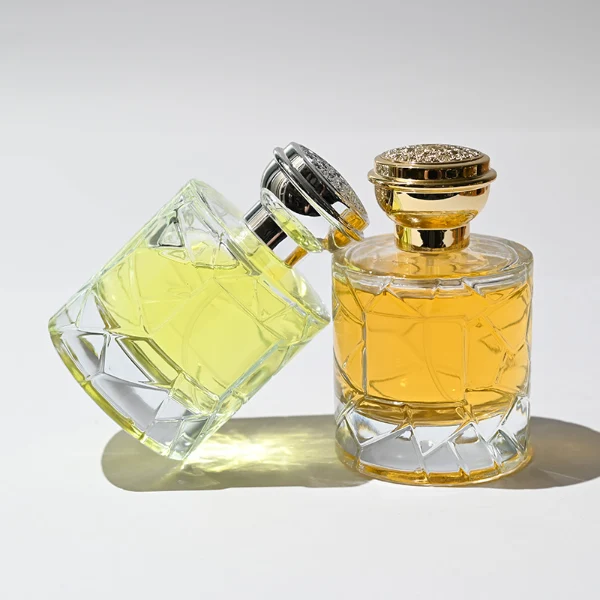HUA0253 perfume bottle, faceted transparent glass body with geometric cut texture and thick base, 100 ml liquid, layered flat cylindrical cap with flat top, narrowed middle section and mirror metallic finish in gold or silver color matching the bottle neck, aluminum label/sticker applied on bottle, modern luxury design, ideal for gifting and daily use.