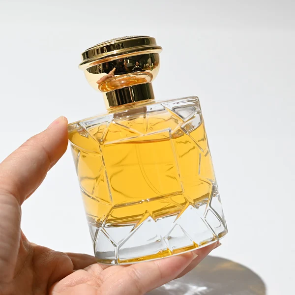 HUA0253 perfume bottle, faceted transparent glass body with geometric cut texture and thick base, 100 ml liquid, layered flat cylindrical cap with flat top, narrowed middle section and mirror metallic finish in gold or silver color matching the bottle neck, aluminum label/sticker applied on bottle, modern luxury design, ideal for gifting and daily use.