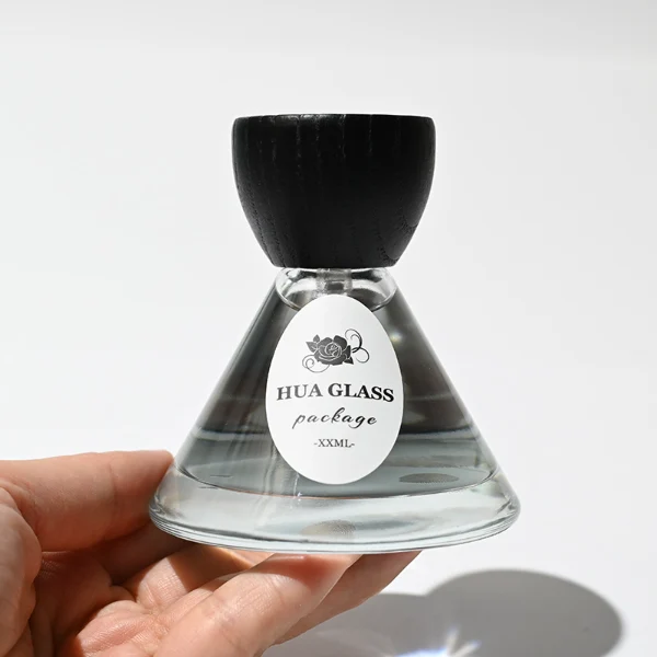 HUA0257 perfume bottle, transparent glass body with wide cone-shaped silhouette and thick rounded base, 30 ml, 50 ml, or 100 ml liquid, rounded wooden cap with matte finish and bowl-shaped silhouette in black or brown color, printed logo label, artistic modern luxury design, ideal for gifting and daily use.