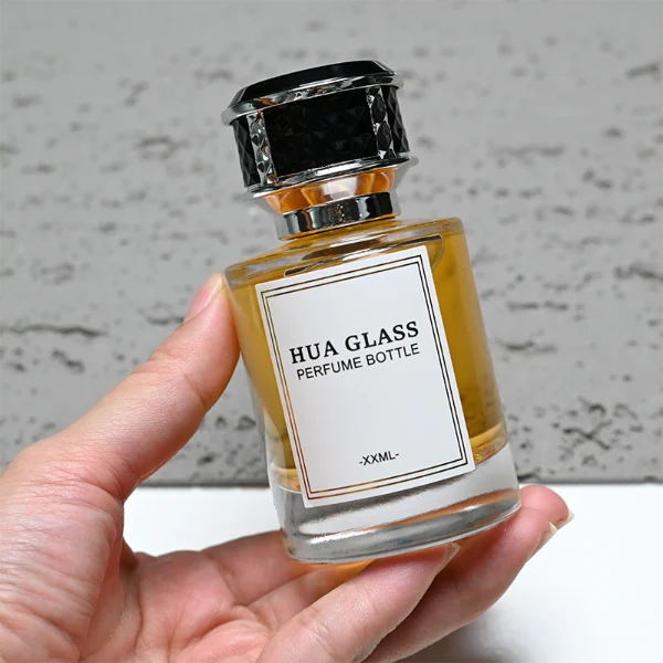 HUA0267 perfume bottle, cylindrical transparent glass body with thick rounded base, 30 ml, 50 ml, or 100 ml liquid, flat cylindrical plastic cap with faceted lattice side texture and gold or silver metallic striped decoration, printed logo label, modern luxury elegance design, ideal for gifting and daily use.