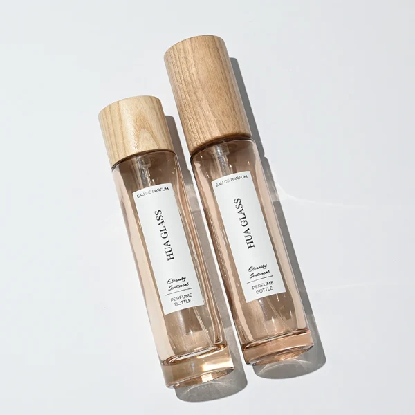 HUA0269 perfume bottle, slim cylindrical transparent glass body with rounded base, 30 ml, 50 ml, or 100 ml liquid, tall cylindrical wooden cap with natural wood grain texture, printed vertical logo label, natural minimalist elegance design, ideal for gifting and travel use.