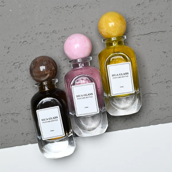 HUA0276 perfume bottle, transparent colorless oval glass body with rounded contours and thick base, 80 ml liquid, glossy spherical resin cap with marble-like texture in customizable colors, printed label attached on bottle surface, modern elegant luxury design, ideal for gifting and daily use.
