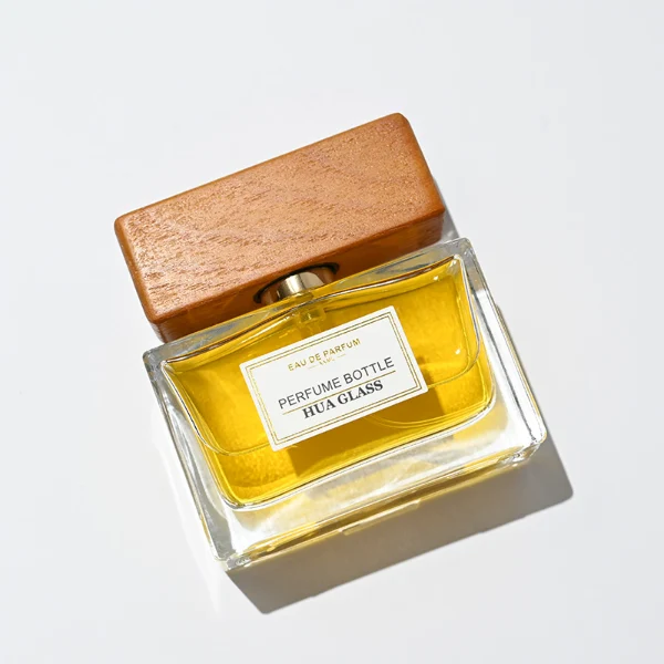 HUA0277 perfume bottle, transparent colorless rectangular glass body with thick base and straight edges, 30 ml, 50 ml, or 100 ml liquid, wide flat rectangular wooden cap in black or natural wood finish with gold bottle neck, printed label attached on bottle surface, modern minimalist luxury design, ideal for gifting and daily use.