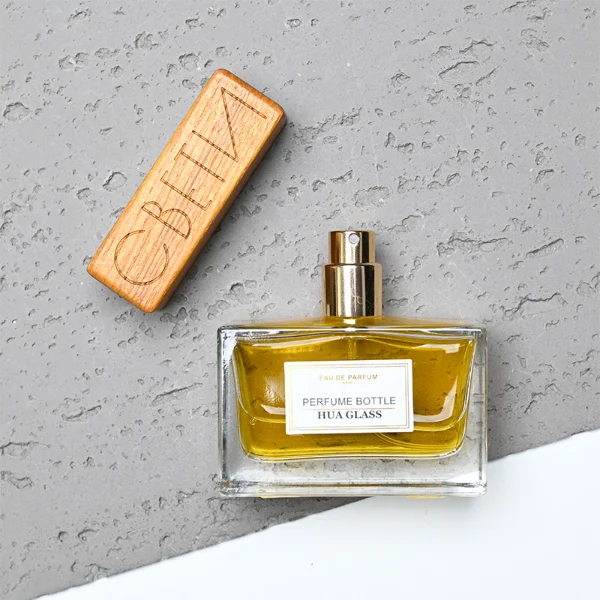 HUA0277 perfume bottle, transparent colorless rectangular glass body with thick base and straight edges, 30 ml, 50 ml, or 100 ml liquid, wide flat rectangular wooden cap in black or natural wood finish with gold bottle neck, printed label attached on bottle surface, modern minimalist luxury design, ideal for gifting and daily use.
