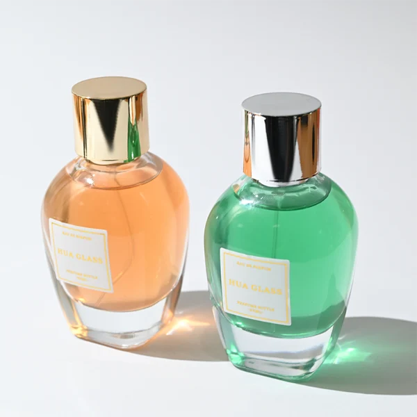 HUA0282 perfume bottle, rounded transparent glass body with thick clear base and customizable transparent colors, 100 ml liquid, glossy cylindrical Zamac cap in silver or gold finish with matching bottle neck, gold-bordered label attached on bottle surface, modern elegant luxury design, ideal for gifting and daily use.