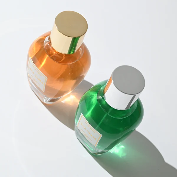 HUA0282 perfume bottle, rounded transparent glass body with thick clear base and customizable transparent colors, 100 ml liquid, glossy cylindrical Zamac cap in silver or gold finish with matching bottle neck, gold-bordered label attached on bottle surface, modern elegant luxury design, ideal for gifting and daily use.
