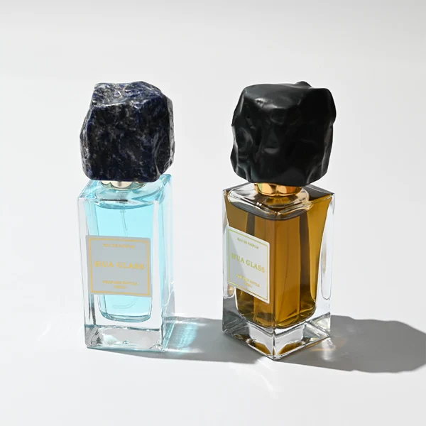 HUA0286 perfume bottle, rectangular transparent colorless glass body with thick base and beveled edges, 30 ml, 50 ml, or 100 ml liquid, irregular natural stone cap with raw textured surface and gold bottle neck, printed label attached on bottle surface, modern artistic luxury design, ideal for gifting and daily use.