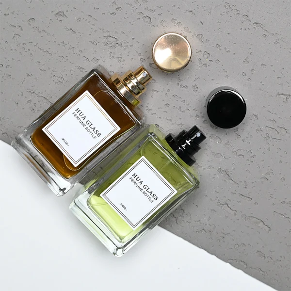 HUA0291 perfume bottle, rectangular transparent colorless glass body with thick base and beveled edges, 100 ml liquid, glossy cylindrical aluminum cap in gold or black finish, printed label attached on bottle surface, modern minimalist luxury design, ideal for gifting and daily use.