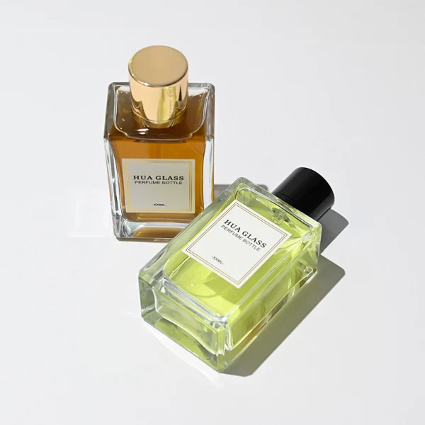 HUA0291 perfume bottle, rectangular transparent colorless glass body with thick base and beveled edges, 100 ml liquid, glossy cylindrical aluminum cap in gold or black finish, printed label attached on bottle surface, modern minimalist luxury design, ideal for gifting and daily use.