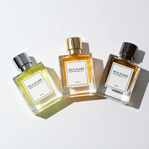 HUA0294 perfume bottle, square transparent colorless glass body with thick base and beveled edges, 50 ml liquid, glossy cylindrical Zamac cap in black, gold, or silver finish with matching bottle neck, printed label attached on bottle surface, modern minimalist luxury design, ideal for gifting and daily use.