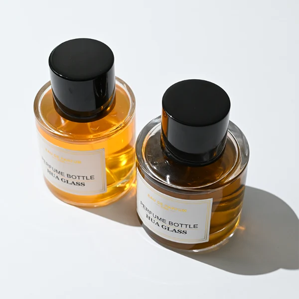 HUA0303 perfume bottle, cylindrical transparent colorless glass body with thick rounded base and smooth curved silhouette, 30 ml, 50 ml, or 100 ml liquid, glossy black cylindrical aluminum cap with flat top, printed label attached on bottle surface, modern minimalist luxury design, ideal for gifting and daily use.