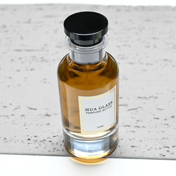 HUA0305 perfume bottle, cylindrical transparent colorless glass body with thick rounded base and curved shoulders, 50 ml or 100 ml liquid, stopper-shaped cylindrical plastic cap in black or white finish, printed label attached on bottle surface, modern minimalist luxury design, ideal for gifting and daily use.