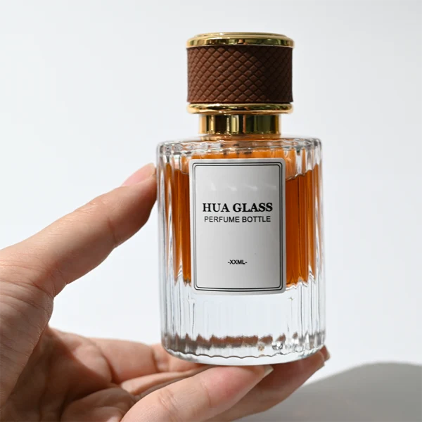 HUA0307 perfume bottle, cylindrical transparent colorless glass body with vertical ribbed texture and thick base, 30 ml, 50 ml, or 100 ml liquid, gold plastic cylindrical cap wrapped with black or brown leather, printed label attached on bottle surface, modern vintage luxury design, ideal for gifting and daily use.