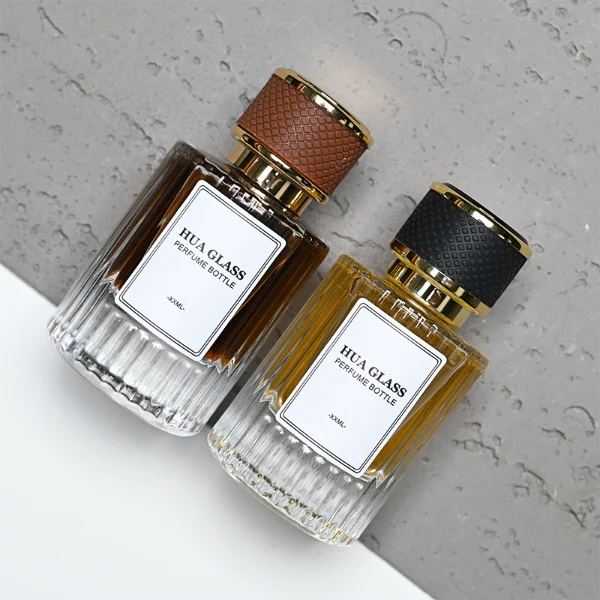 HUA0307 perfume bottle, cylindrical transparent colorless glass body with vertical ribbed texture and thick base, 30 ml, 50 ml, or 100 ml liquid, gold plastic cylindrical cap wrapped with black or brown leather, printed label attached on bottle surface, modern vintage luxury design, ideal for gifting and daily use.