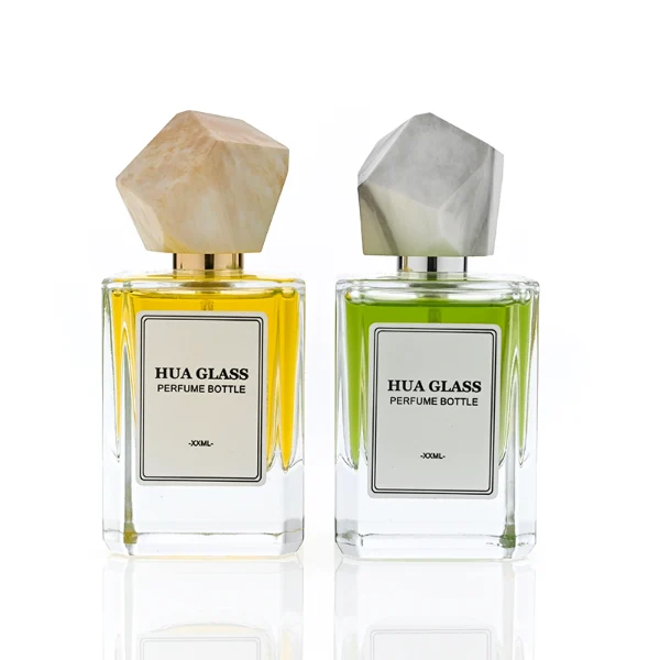 HUA0322 perfume bottle, square transparent colorless glass body with thick base and sharp edges, 30 ml, 50 ml, or 100 ml liquid, irregular geometric stone-inspired plastic cap in beige or marble white color with gold or silver bottle neck, printed label attached on bottle surface, artistic modern luxury design, ideal for gifting and daily use.