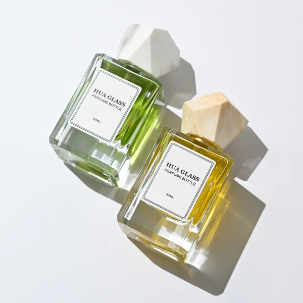 HUA0322 perfume bottle, square transparent colorless glass body with thick base and sharp edges, 30 ml, 50 ml, or 100 ml liquid, irregular geometric stone-inspired plastic cap in beige or marble white color with gold or silver bottle neck, printed label attached on bottle surface, artistic modern luxury design, ideal for gifting and daily use.