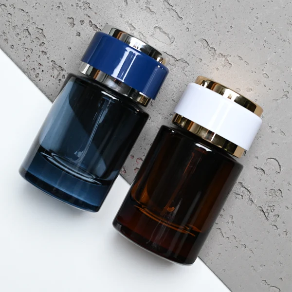 HUA0326 perfume bottle, rounded cylindrical semi-transparent smoky black, amber brown, or blue glass body with thick base, 30 ml, 50 ml, or 100 ml liquid, layered cylindrical plastic cap with black, white, or blue center section and glossy gold or silver accents on both ends, optional metallic gold printed logo or blank bottle surface, modern minimalist luxury design, ideal for gifting and daily use.