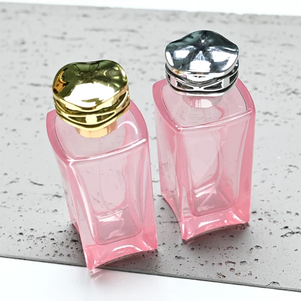 HUA0338 perfume bottle, rectangular transparent pink glass body with curved inner contour and thick base, 100 ml liquid, irregular geometric plastic cap with wave-pattern side texture in gold or silver finish, matching metallic bottle neck, clean bottle surface for logo printing or label application, elegant feminine luxury design, ideal for gifting and daily use.