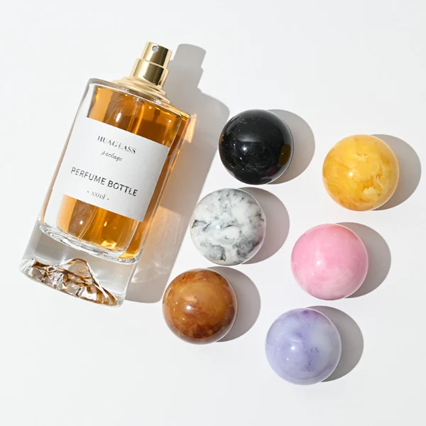 HUA0064 perfume bottle, heavy-base cylindrical round glass bottle with clear finish and mountain-shaped inner base, featuring a square paper label, a matching neck, and a playful spherical ball-shaped synthetic polymer cap with an organic marbled or solid polished finish, elegant modern fragrance packaging design available in 30ml, 50ml, and 100ml