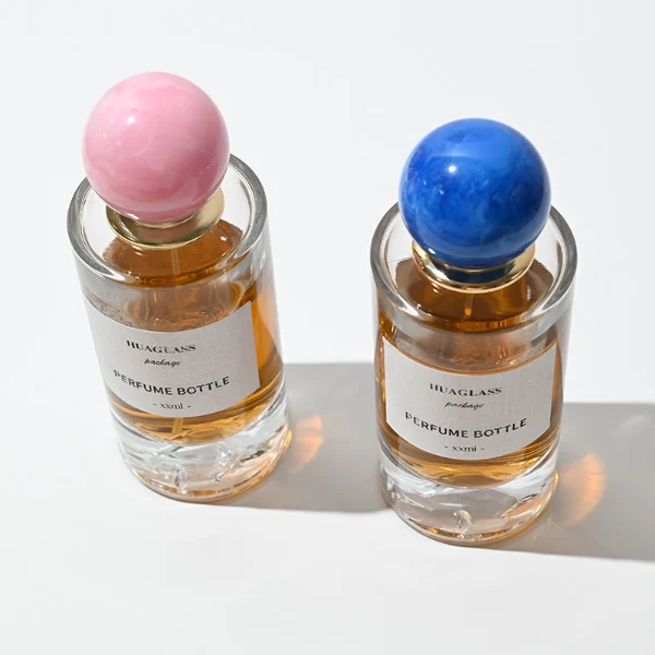 HUA0064 perfume bottle, heavy-base cylindrical round glass bottle with clear finish and mountain-shaped inner base, featuring a square paper label, a matching neck, and a playful spherical ball-shaped synthetic polymer cap with an organic marbled or solid polished finish, elegant modern fragrance packaging design available in 30ml, 50ml, and 100ml