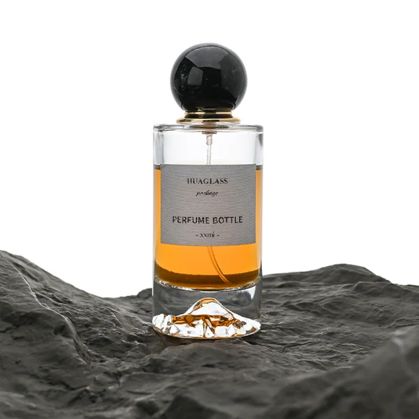 HUA0064 perfume bottle, heavy-base cylindrical round glass bottle with clear finish and mountain-shaped inner base, featuring a square paper label, a matching neck, and a playful spherical ball-shaped synthetic polymer cap with an organic marbled or solid polished finish, elegant modern fragrance packaging design available in 30ml, 50ml, and 100ml