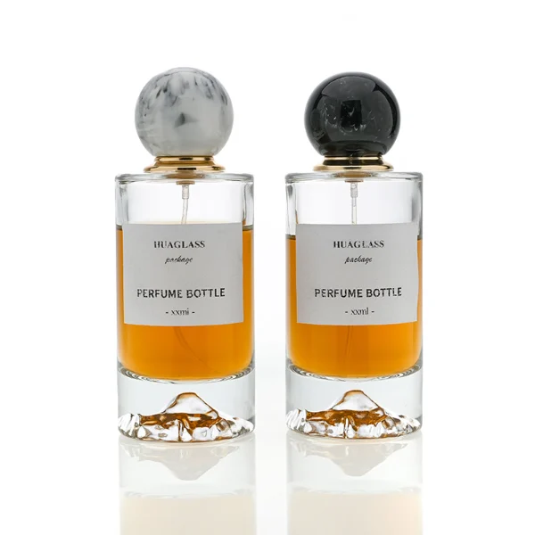HUA0064 perfume bottle, heavy-base cylindrical round glass bottle with clear finish and mountain-shaped inner base, featuring a square paper label, a matching neck, and a playful spherical ball-shaped synthetic polymer cap with an organic marbled or solid polished finish, elegant modern fragrance packaging design available in 30ml, 50ml, and 100ml
