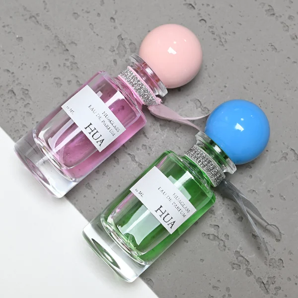 HUA0063 perfume bottle, heavy-base slender cylindrical round glass bottle with clear finish, featuring a square paper label, a matching neck with ribbon bow decorations, and a playful spherical ball-shaped plastic cap with a smooth high-gloss shiny finish, elegant modern fragrance packaging design available in 30ml, 50ml, and 100ml