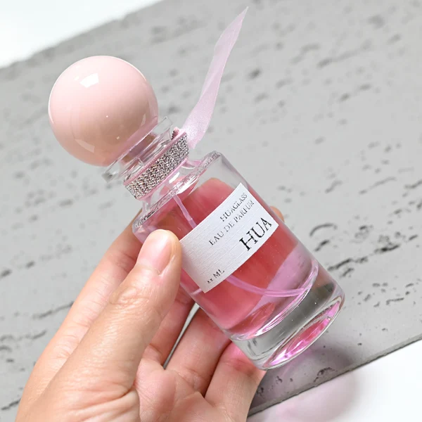 HUA0063 perfume bottle, heavy-base slender cylindrical round glass bottle with clear finish, featuring a square paper label, a matching neck with ribbon bow decorations, and a playful spherical ball-shaped plastic cap with a smooth high-gloss shiny finish, elegant modern fragrance packaging design available in 30ml, 50ml, and 100ml