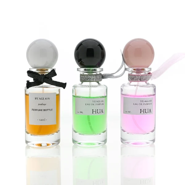 HUA0063 perfume bottle, heavy-base slender cylindrical round glass bottle with clear finish, featuring a square paper label, a matching neck with ribbon bow decorations, and a playful spherical ball-shaped plastic cap with a smooth high-gloss shiny finish, elegant modern fragrance packaging design available in 30ml, 50ml, and 100ml