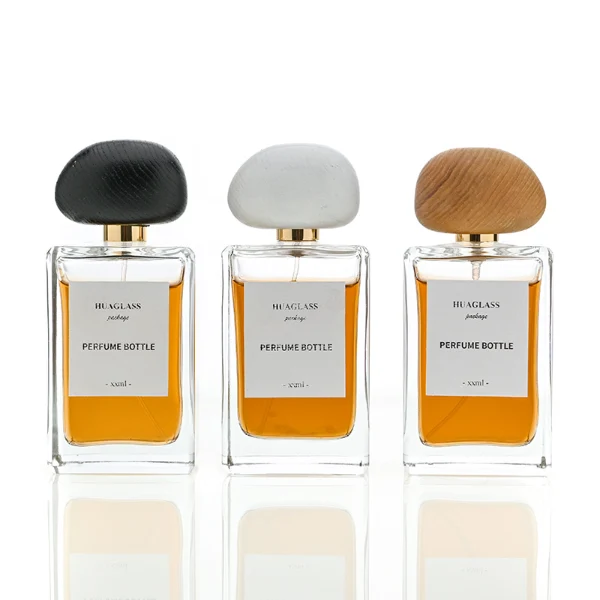 HUA0014 perfume bottle, heavy-base rectangular cuboid glass bottle with clear finish, featuring a square paper label, a polished gold neck, and an organic pebble-shaped timber cap with a natural smooth grain finish, elegant modern fragrance packaging design available in 100ml