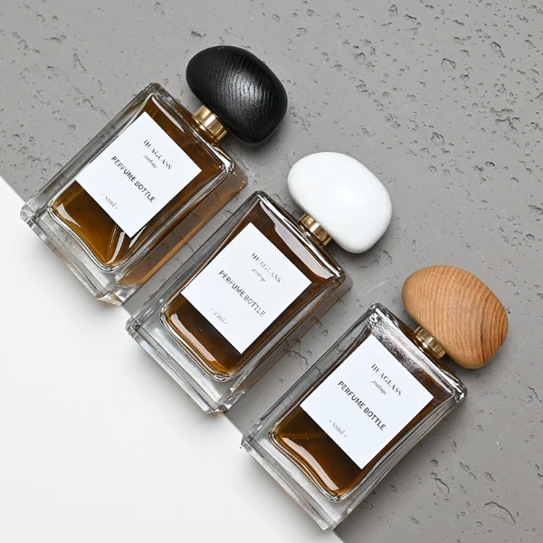 HUA0014 perfume bottle, heavy-base rectangular cuboid glass bottle with clear finish, featuring a square paper label, a polished gold neck, and an organic pebble-shaped timber cap with a natural smooth grain finish, elegant modern fragrance packaging design available in 100ml