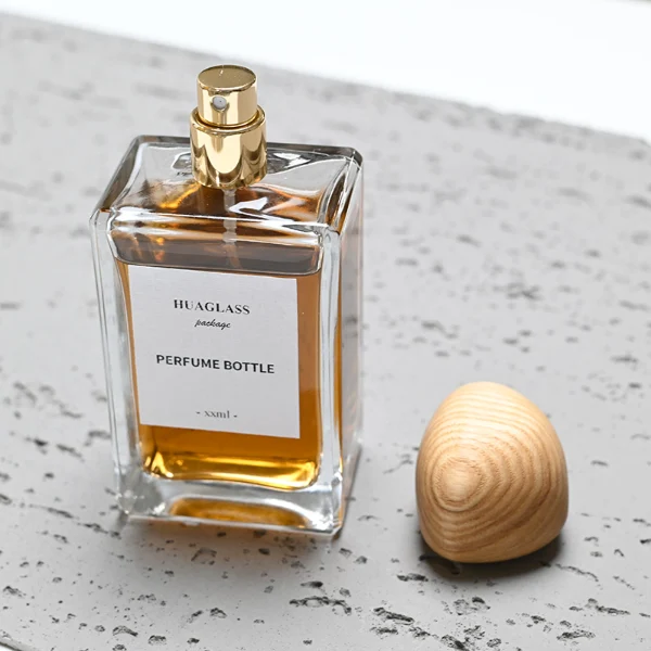 HUA0014 perfume bottle, heavy-base rectangular cuboid glass bottle with clear finish, featuring a square paper label, a polished gold neck, and an organic pebble-shaped timber cap with a natural smooth grain finish, elegant modern fragrance packaging design available in 100ml