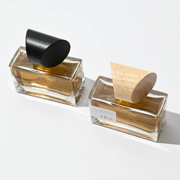 HUA0054 perfume bottle, heavy-base wide rectangular cuboid glass bottle with clear finish, featuring a gold collar neck accent and a unique horizontally oriented cylindrical barrel-shaped wood cap with subtle grain textures in a shiny charcoal black or natural light wood finish, elegant modern fragrance packaging design available in 30ml, 50ml, and 100ml.
