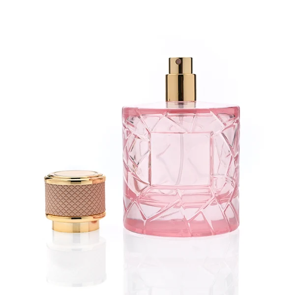 HUA0342 perfume bottle, cylindrical transparent pink glass body with fragmented surface texture, 50 ml or 100 ml liquid, gold plastic cap with textured pink or black side surface and glossy gold top, gold bottle neck, aluminum brand label attached on bottle surface, modern feminine luxury design, ideal for gifting and daily use.