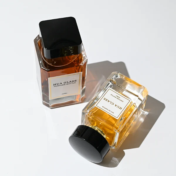 HUA0344 perfume bottle, rectangular transparent colorless glass body with beveled edges and thick base, 30 ml, 50 ml, or 100 ml liquid, wide glossy black cylindrical plastic cap with convex top and flat sides, gold screw-type bottle neck, brand logo label attached on bottle surface, modern minimalist luxury design, ideal for gifting and daily use.