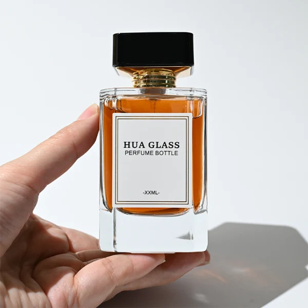 HUA0344 perfume bottle, rectangular transparent colorless glass body with beveled edges and thick base, 30 ml, 50 ml, or 100 ml liquid, wide glossy black cylindrical plastic cap with convex top and flat sides, gold screw-type bottle neck, brand logo label attached on bottle surface, modern minimalist luxury design, ideal for gifting and daily use.
