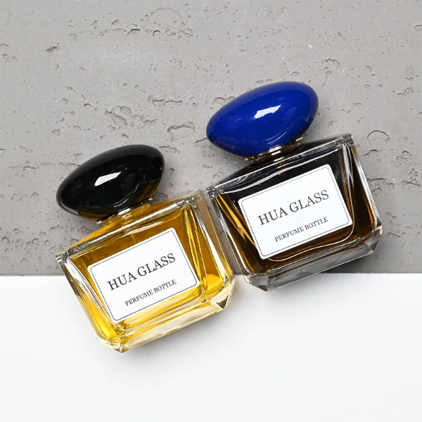 HUA0359 perfume bottle, low rectangular clear glass body with gold neck and a paper label attached, smooth oval pebble plastic cap in navy blue, white, or burgundy, contemporary artistic luxury fragrance packaging design available in 50ml and 100ml