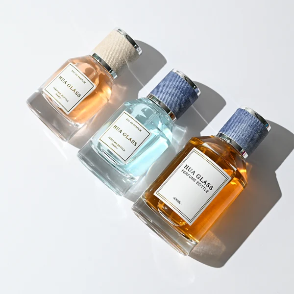 HUA0365 perfume bottle, cylindrical clear glass body with faceted sloped shoulders, silver neck and a paper label attached, textured blue or beige cylindrical plastic cap with silver trim, contemporary casual luxury fragrance packaging design available in 30ml, 50ml, and 100ml